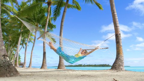 LOW ANGLE: Female traveler unwinds by the breathtaking ocean by taking an afternoon nap in a rope hammock. Relaxed woman enjoying her summer vacation on tropical island by swaying under the palm trees