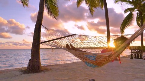 SLOW MOTION, LENS FLARE, CLOSE UP: Carefree girl napping in a swaying hammock on golden summer evening on the tranquil sandy beach in Cook Islands. Female tourist relaxing on the beach at sunrise.