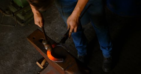 Overhead of young female metalsmith with short hair molding horseshoe in factory. She hit the glowing warm horseshoe with a hammer.