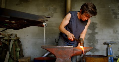 Attentive female metalsmith molding a glowing hot horseshoe in factory, hitting it with a hammer.