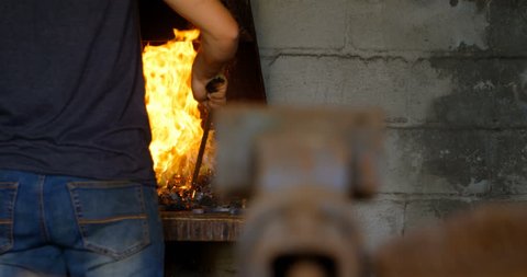 Rear view of a young female metalsmith heating horseshoe in fire at factory. Looking over the shoulder, smiling at the camera at the end.