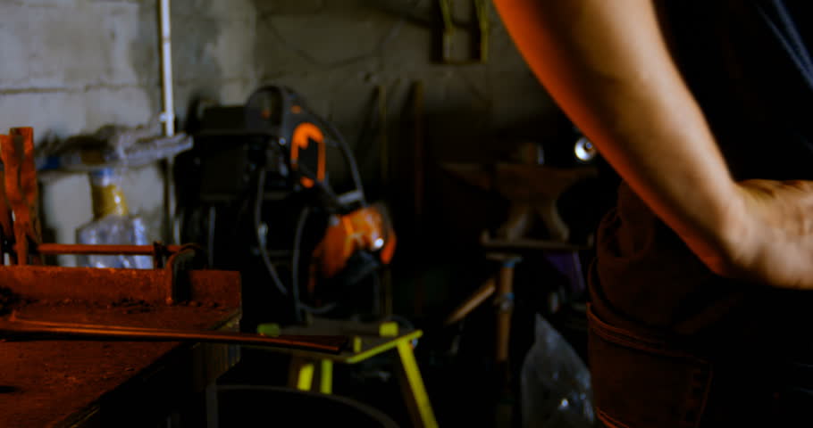 Side view of female metalsmith standing in factory wearing safety glasses. Swiping the sweat of her forehead.