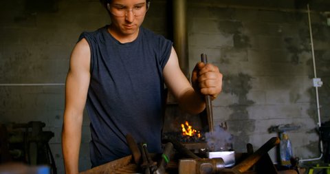 Front view of young female metalsmith with short hair working in workshop. Fire in the background.