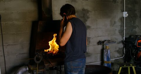 Rear view of female metalsmith talking on mobile phone while working in factory in front of the fire.