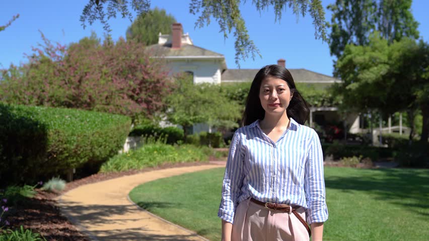 focus on elegant female household standing outdoors raising her hand showing house with beautiful green garden smiling to camera. confident asian woman in sunlight on summer with blue clean sky. Royalty-Free Stock Footage #1020848698