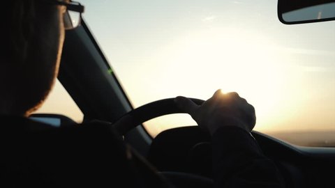 A man rides his car in the rays of sunset. Reflection in the rearview mirror. Man hands holding Steering wheel while driving car. Hands of a driver on steering wheel of a car and empty asphalt road