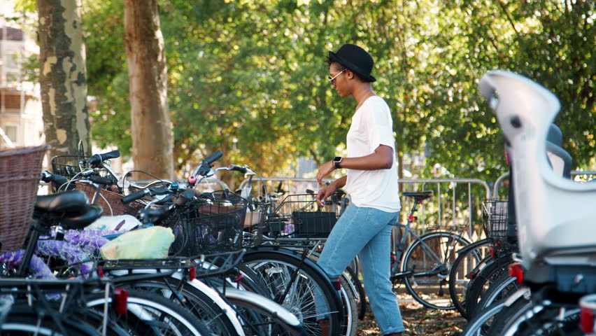Young black woman wearing white t shirt, blue jeans and sunglasses parking her bicycle, putting on crossbody handbag and walking away smiling, handheld
