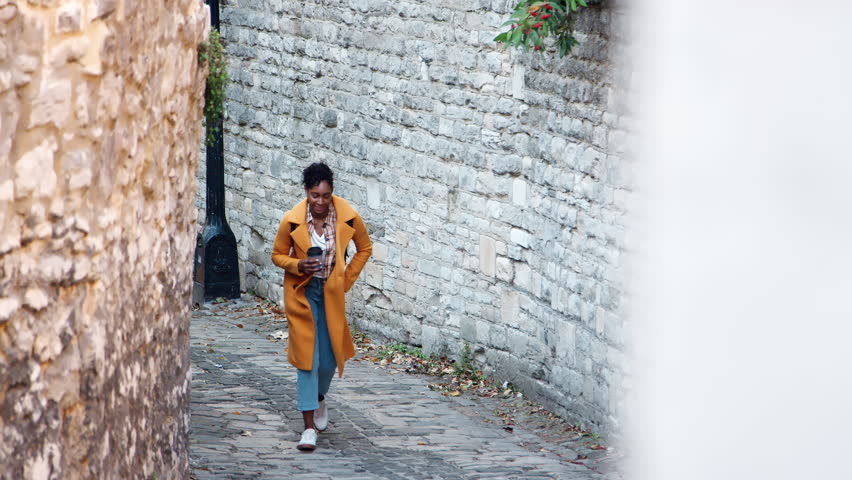 Front view of young black woman wearing a yellow pea coat holding a takeaway coffee walking towards camera in an alleyway between stone walls, elevated view