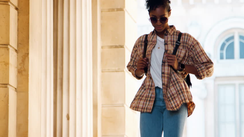 Close up of a fashionable young black woman wearing sunglasses and a plaid shirt walking down stairs outside a historical building, selective focus