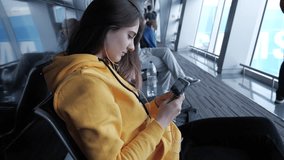 Woman wait for boarding in airport, spend time by watching film on smartphone. Girl with headphones stare to mobile device screen