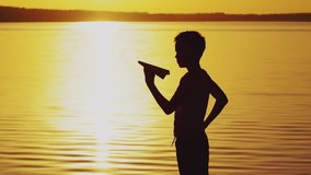 A little boy is holding a paper airplane in his hand and playing with him on the bank of the river at sunset on the background of the solar track.