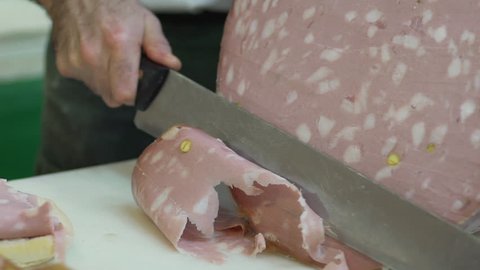 Butcher slicing organic Italian Bologna sausage mortadella, a traditional pork sausage, with a sharp knife to prepare a delicious ban. Buns with mortadella, food. Slow motion, 