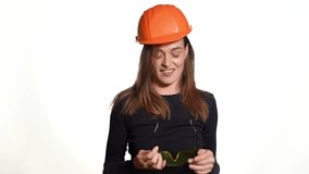 A joyful woman in orange helmet for building is trying on goggles and posing in it on a white background in the studio.