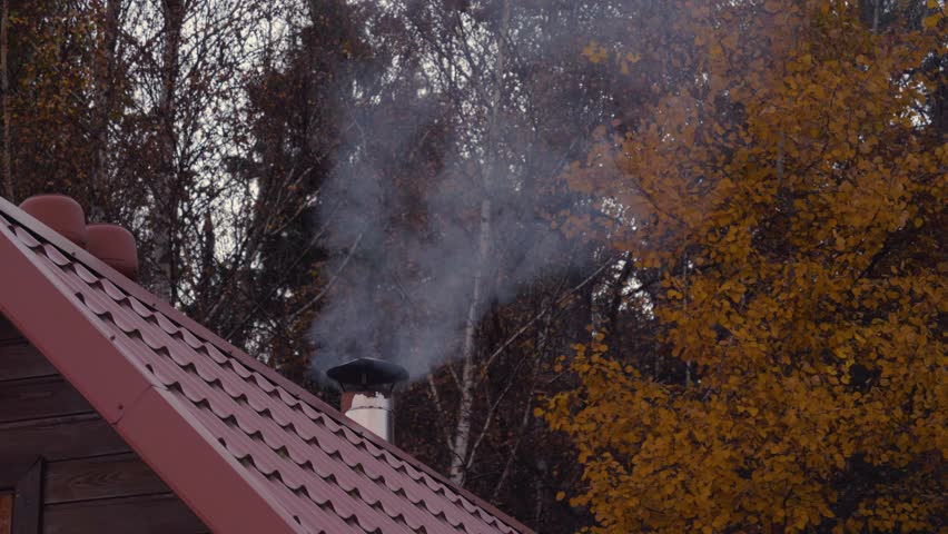 White smoke comes from the chimney of the house and rises into the air, house with a red tiled roof on a cold autumn evening, against the forest with yellow foliage