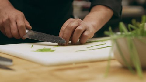 Focused professional chef using a knife to thinly slice herbs on a cutting board in industrial kitchen with soft interior lighting. Close up shot on 4k RED camera.