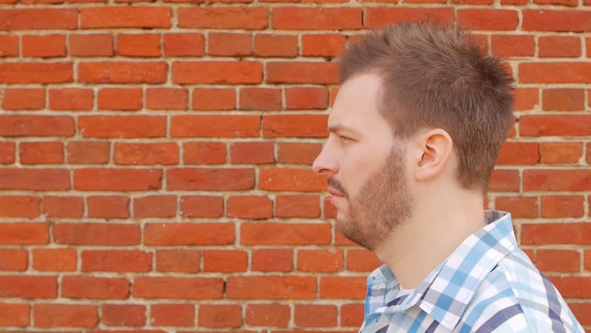 Handsome stylish man smoking vap against a brick wall and looking into the camera, slow-mo, fashionable
