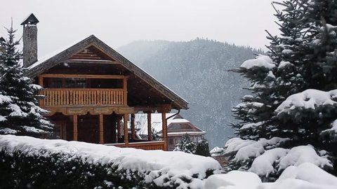 Christmas wooden mansion in mountains on snowfall winter day. Cozy chalet on ski resort near pine forest. Cottage of round timber with wooden balcony. Fir-trees covered with snow. Chimneys of stone.