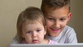 Two children watch video on laptop and have fun together at home. Big brother and small sister raptly look to screen, learn and laugh. Siblings, boy and toddler girl smile looking to gadget display