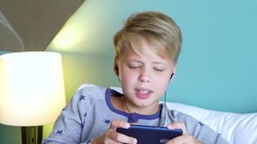 Closeup portrait of cute preteen kid listening to music and singing. Boy laying in bed and holding modern mobile smartphone in hands. Real time 4k video footage.