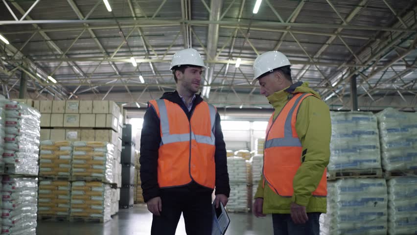 Two employees in workwear and white helmets discussing work in warehouse. Royalty-Free Stock Footage #1020927790