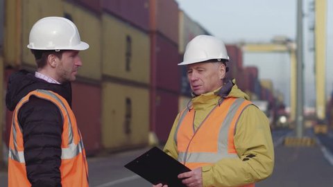 Port manager and a colleague tracking inventory while standing together by freight containers on a large commercial shipping dock