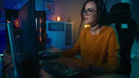 Excited and Concentrated Gamer Girl in Glasses and Headset with a Mic Playing Online Strategy Video Game on Her Personal Computer. Room and PC have Colorful Warm Neon Led Lights. Cozy Evening at Home.