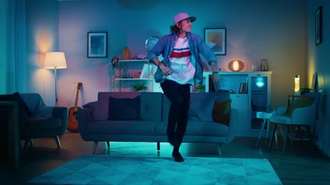 Handsome Excited Hip Young Man is Dancing in the Living Room while TV Plays in the Background. He is Energetically Moving while Screen Adds Reflections on Him. Cozy Room is Lit with Warm Neon Light.