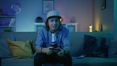 Handsome Excited Young Gamer with Long Hair and a Cap is Sitting on a Couch, Playing and Winning in Video Games on Console. He Plays with a Wireless Controller. Cozy Room with Warm and Neon Light.