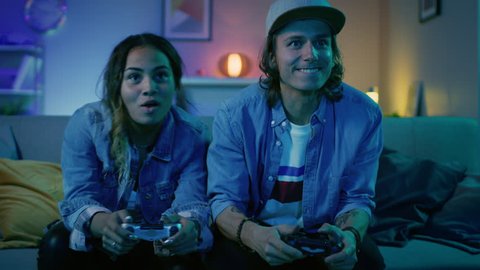 Excited Black Gamer Girl and Young Man Sitting on a Couch and Playing Video Games on Console. They Plays with Wireless Controllers. Cozy Room is Lit with Warm and Neon Light.
