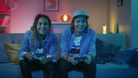 Excited Black Gamer Girl and Young Man Sitting on a Couch and Playing Video Games on Console. They Plays with Wireless Controllers and Give High Five. Cozy Room is Lit with Warm and Neon Light.
