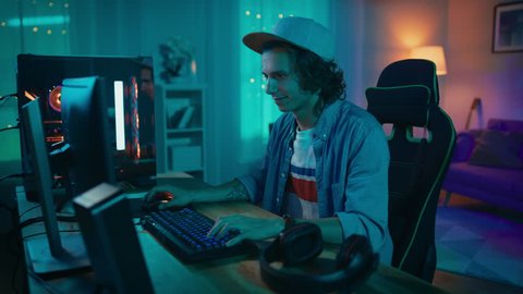 Excited Gamer Playing and Winning in Online Video Game on His Personal Computer. Room and PC have Colorful Warm Neon Led Lights. Young Man is Wearing a Cap. Cozy Evening at Home.