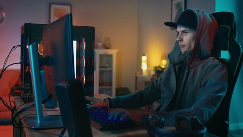 Professional Gamer Playing First-Person Shooter Online Video Game on His Cool Personal Computer. Young Man is Wearing a Cap and Hood. Room and PC have Colorful Neon Led Lights. Cozy Evening at Home.