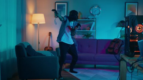 Young Man is Energetically Playing a Virtual Reality Video Game While Wearing a Headset. He's is Standing in a Cozy Room with Warm Neon Lights. He's Swinging Hands, Shooting and Dodging Obstacles.