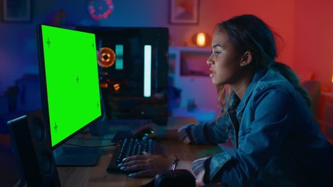 Beautiful Black Girl Freelance Designer Works on a Powerful Gamers Computer with a Green Screen Mock Up at Home. She's Concentrated. Cozy Room is Lit with Warm and Neon Light.