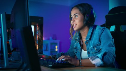Pretty and Excited Black Gamer Girl in Headphones is Playing and Winning in First-Person Shooter Online Video Game on Her Computer. Room and PC have Colorful Neon Led Lights. Cozy Evening at Home.