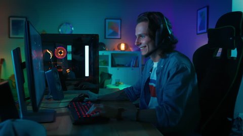 Excited Gamer Comes to the Table, Puts On His Headset with a Mic and Starts Playing Online Video Game on His Personal Computer. Room and PC have Colorful Neon Led Lights. Cozy Evening at Home.