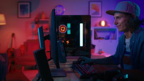 Excited Gamer Playing First-Person Shooter Online Video Game on His Personal Computer. Room and PC have Colorful Neon Led Lights. Young Man is Wearing a Cap. Cozy Evening at Home.