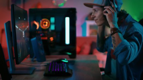 Gamer Puts His Headset with a Mic On and Starts Playing Shooter Online Video Game on His Personal Computer. Room and PC have Colorful Neon Led Lights. Young Man is Wearing a Cap. Cozy Evening at Home.