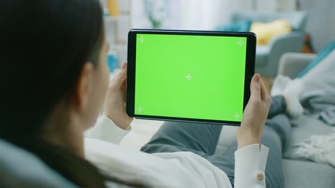 Young Woman at Home Resting on a Couch Using with Green Mock-up Screen Tablet Computer in Horizontal Landscape Portrait Mode. Woman Using Tablet Device, Browsing Internet, Watching Content, Videos.