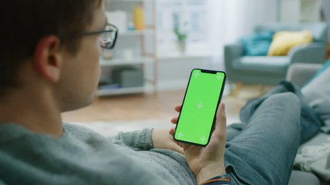 Man at Home Resting on a Couch using Smartphone with Green Mock-up Screen, Doing Swiping, Scrolling Gestures. Guy Using Mobile Phone, Internet Social Networks Browsing.