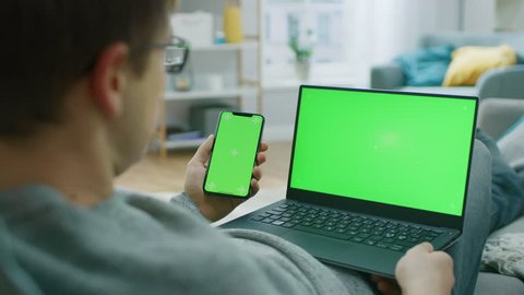 Young Man at Home Works on a Laptop Computer with Green Mock-up Screen, while Holding Smartphone with Chroma Key Display. He's Sitting On a Couch in His Cozy Living Room. Over the Shoulder Camera Shot