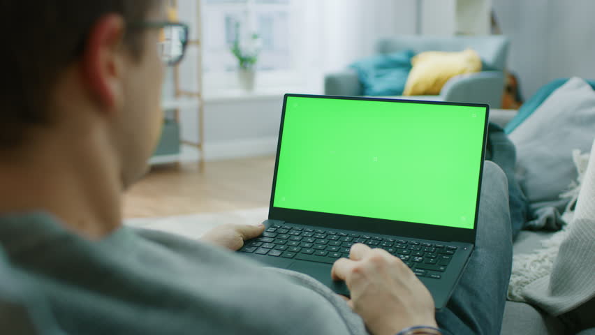 Man at Home Sitting on a Couch Working on Green Mock-up Screen Laptop Computer. Guy Using Laptop Device, Browsing Internet, Watching Content, Videos. Over the Shoulder Camera Shot. Royalty-Free Stock Footage #1020934117