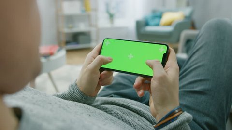 Man at Home Lying on a Couch using Smartphone, Holds it Horizontally in Landscape Mode. Pushing Buttons to Play Video Game. Screen Has Tracking Markers. Point of View Camera Shot.