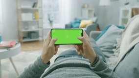 Man at Home Lying on a Couch using Smartphone, Holds it Horizontally in Landscape Mode. Pushing Buttons to Play Video Game. Screen Has Tracking Markers. Point of View Camera Shot.