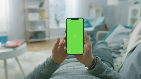 Man at Home Lying on a Couch using Smartphone with Green Mock-up Screen, Doing Swiping, Scrolling Gestures. Guy Using Mobile Phone, Internet Social Networks Browsing. Point of View Camera Shot.
