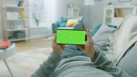 Man at Home Lying on a Couch using Smartphone, Holds it Horizontally in Landscape Mode. Playing Video Games, Watching Videos. Screen Has Tracking Markers. Point of View Camera Shot.