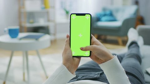Woman at Home Lying on a Couch using Smartphone with Green Mock-up Screen, Doing Swiping, Scrolling Gestures. Girl Using Mobile Phone, Internet Social Networks Browsing. Point of View Camera Shot.