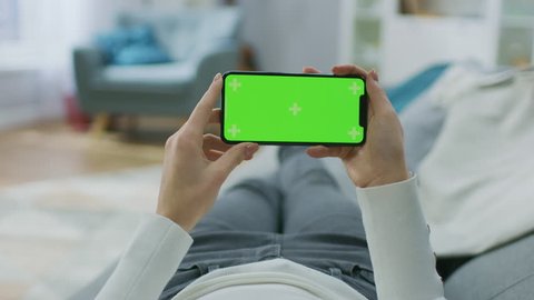 Young Woman at Home Lying on a Couch using with Green Mock-up Screen Smartphone in Horizontal Landscape Mode. Girl Using Mobile Phone, Browsing Internet, Watching Content, Videos, Blogs. POV.