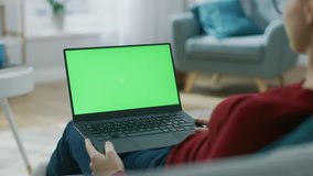 Young Woman at Home Works on a Laptop Computer with Green Mock-up Screen. She's Sitting On a Couch in His Cozy Living Room. Over the Shoulder Camera Shot. Shot on 8K RED Camera.