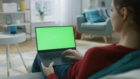Young Woman at Home Works on a Laptop Computer with Green Mock-up Screen. She's Sitting On a Couch in His Cozy Living Room. Over the Shoulder Camera Shot. Shot on 8K RED Camera.